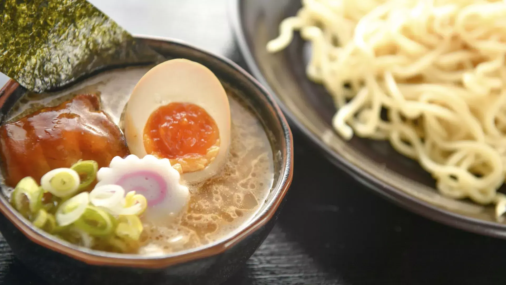 Nahaufnahme einer Schüssel Nudeln und einer Schüssel Ramen-Suppe mit einem pochierten Ei, in zwei Hälften geschnitten.